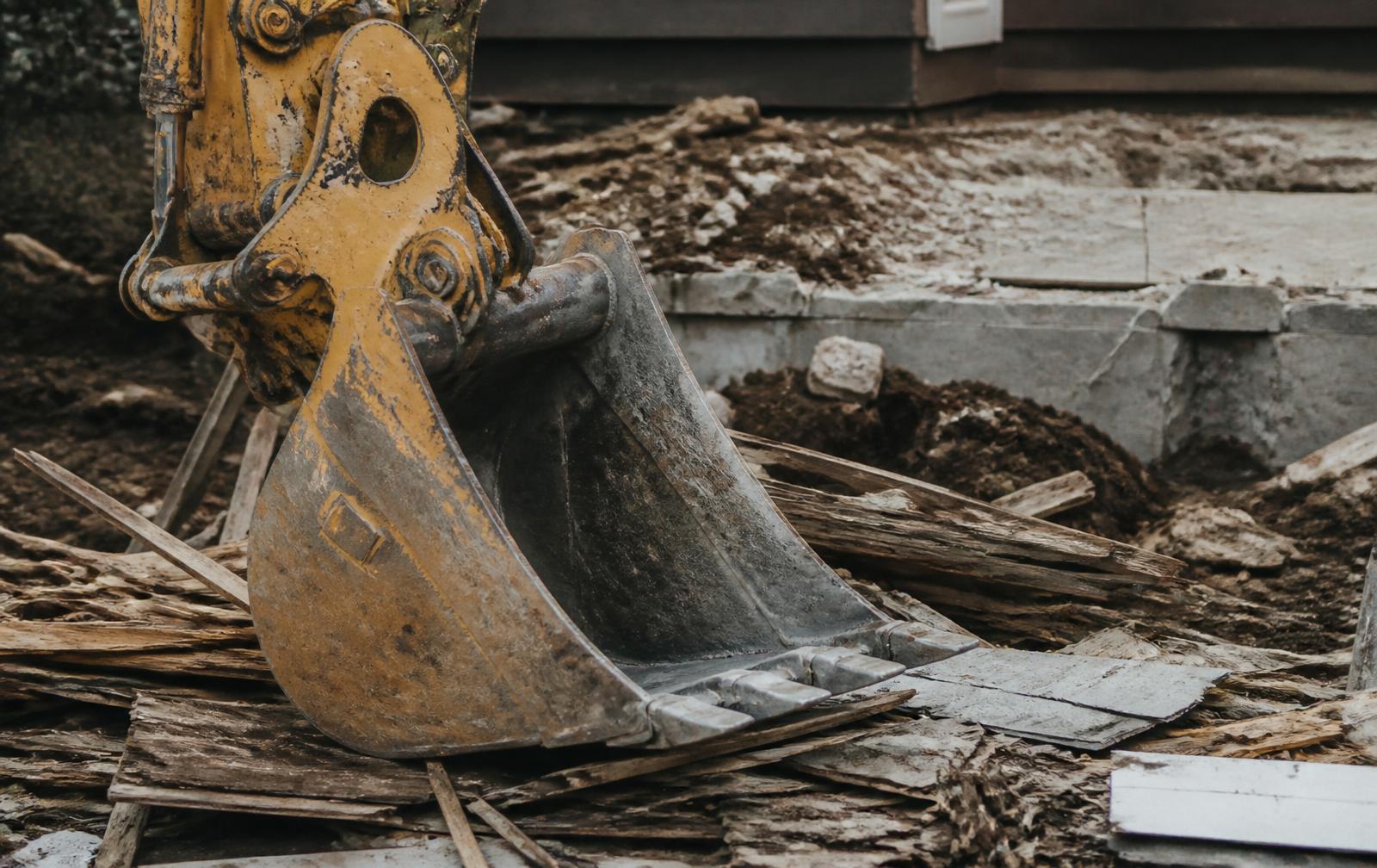 Weathered excavator bucket resting on splintered wood siding and torn shingles on a jobsite